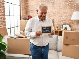 Ein älterer Mann steht in einem Zimmer mit Umzugskartons und hält ein "NEW HOME"-Schild in der Hand, nachdem er mit Hilfe von A2B Umzüge in sein neues Zuhause umgezogen ist.