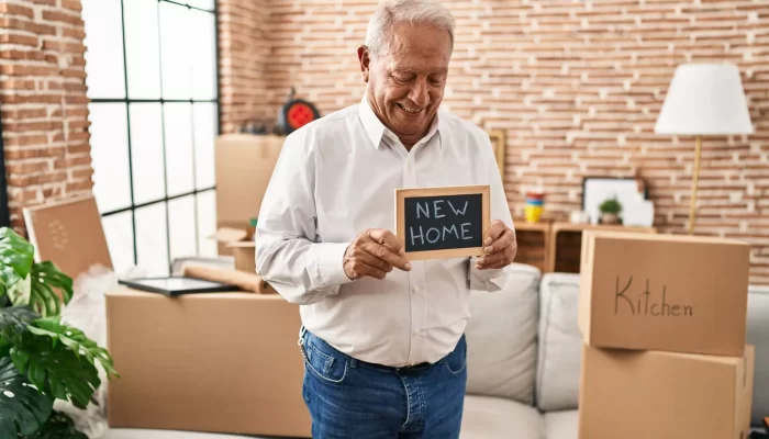 Ein älterer Mann steht in einem Zimmer mit Umzugskartons und hält ein "NEW HOME"-Schild in der Hand, nachdem er mit Hilfe von A2B Umzüge in sein neues Zuhause umgezogen ist.