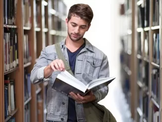 Ein junger Mann blättert in einem Buch und steht zwischen den Regalen einer Bibliothek, nachdem er mit A2B Umzüge in sein neues Zuhause gezogen ist.