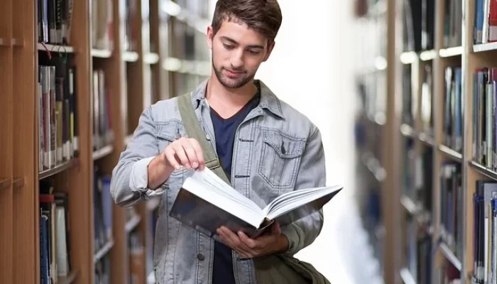 Ein junger Mann blättert in einem Buch und steht zwischen den Regalen einer Bibliothek, nachdem er mit A2B Umzüge in sein neues Zuhause gezogen ist.